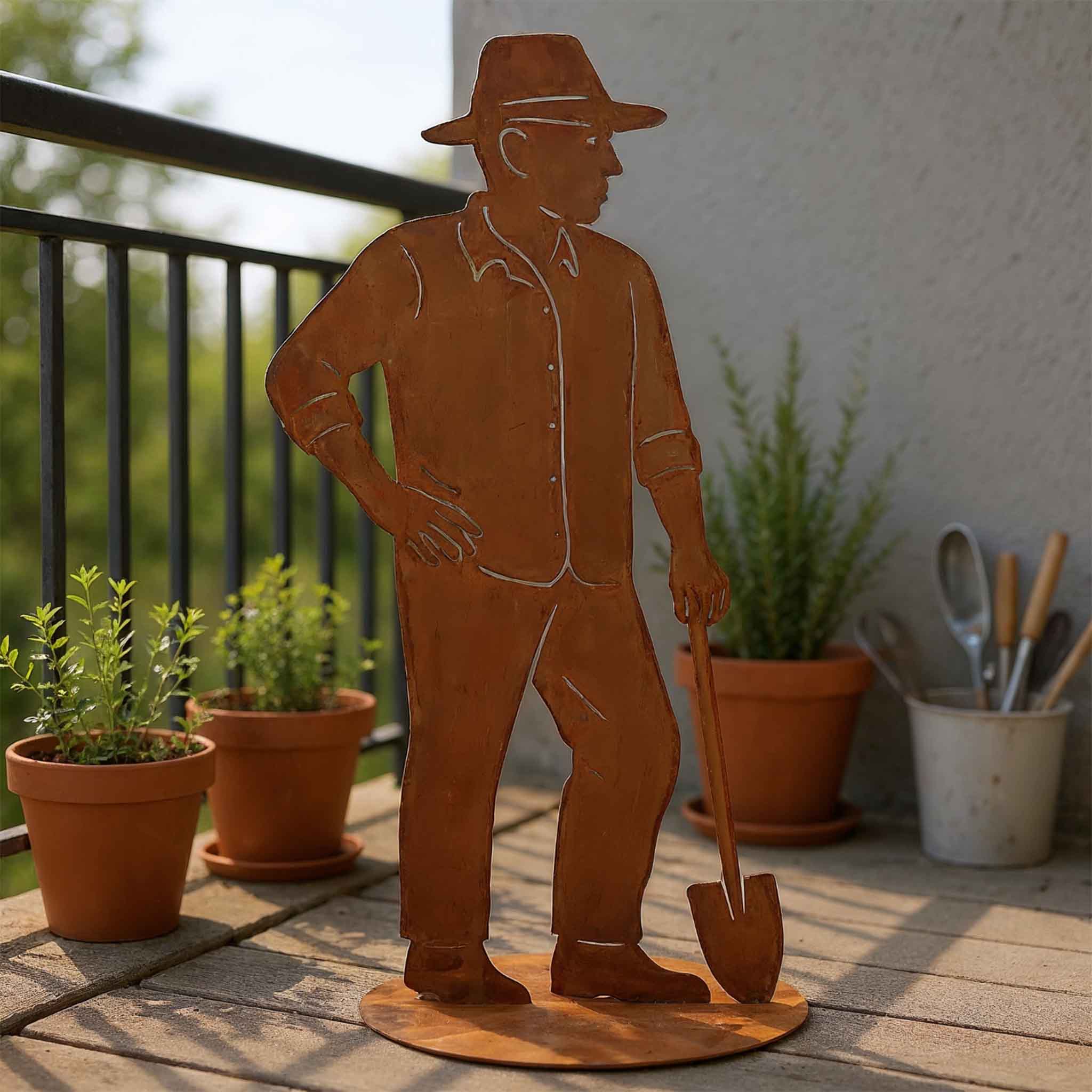Edelrost Gärtner Figur aus Metall mit Schaufel, auf runder Bodenplatte auf einem Balkon aufgestellt. Dekorative Gartendeko mit Kräutertöpfen im Hintergrund.