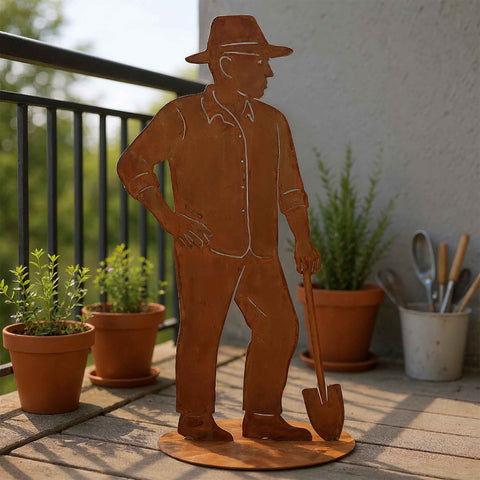 Edelrost Gärtner Figur aus Metall mit Schaufel, auf runder Bodenplatte auf einem Balkon aufgestellt. Dekorative Gartendeko mit Kräutertöpfen im Hintergrund.