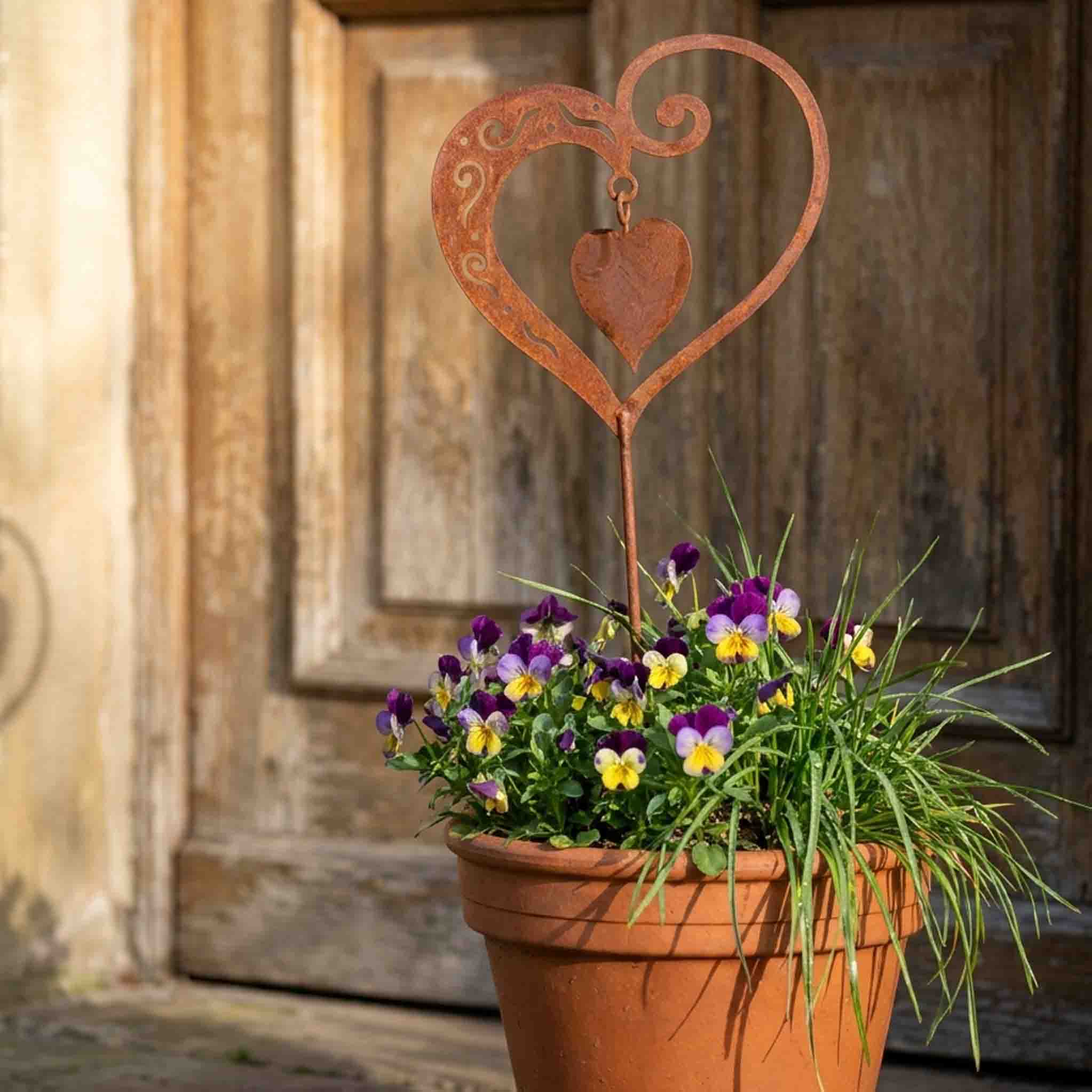 Edelrost Herz mit beweglichem Innenherz als Gartenstecker im Terracotta-Topf mit Hornveilchen vor rustikaler Holztür - handgefertigt in Bayern von Rostikal